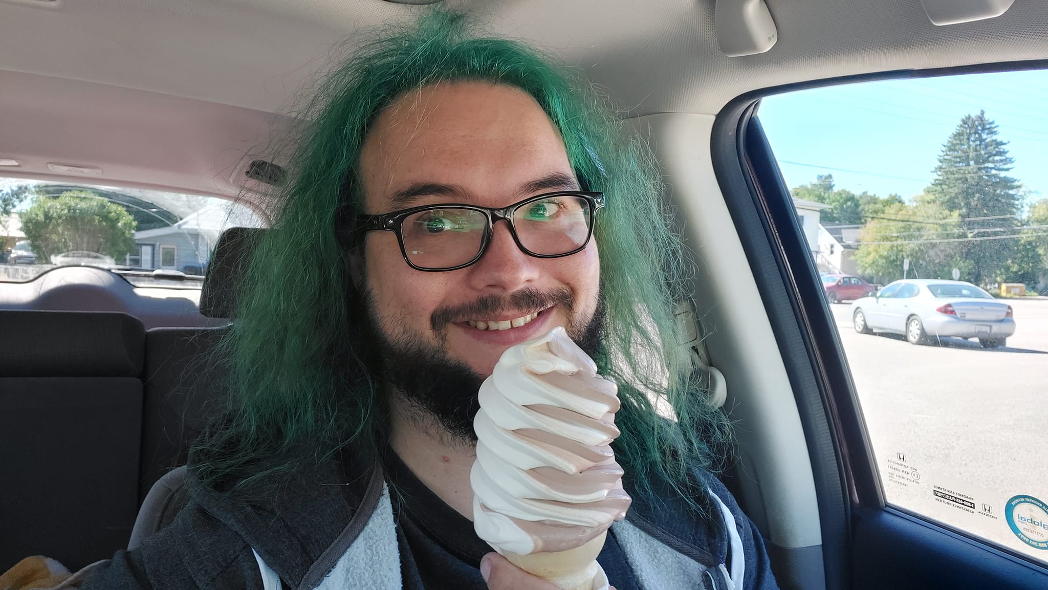 A man with green hair is sitting in a car with a swirrled soft serve ice cream cone